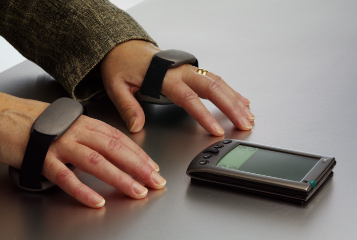 Dwie dłonie spoczywające obok palmtopa, na śródręczu każdej urządzenia przypominające ręczny zegarek